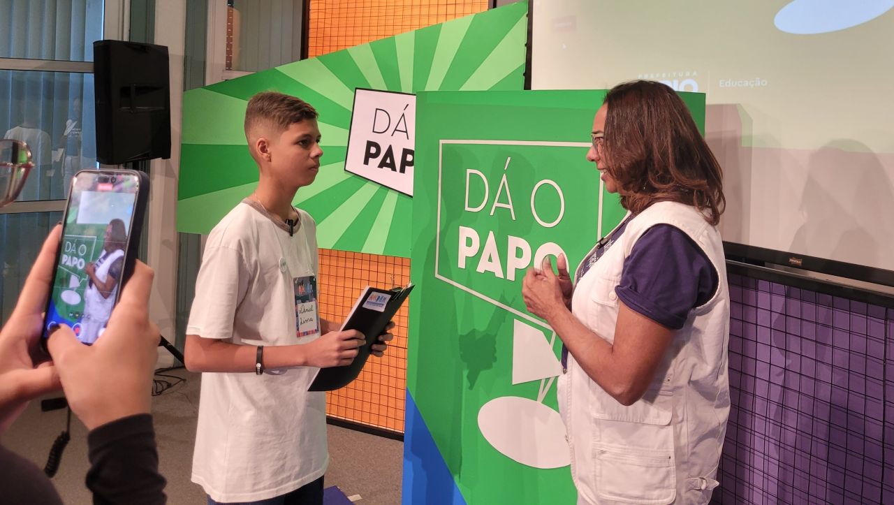 Foto. Um jovem estudante entrevista uma mulher adulta. No fundo, painéis coloridos, com o logotipo do jogo Dá o papo. Em primeiro plano, no canto esquerdo, uma pessoa segura um celular registrando a entrevista.
