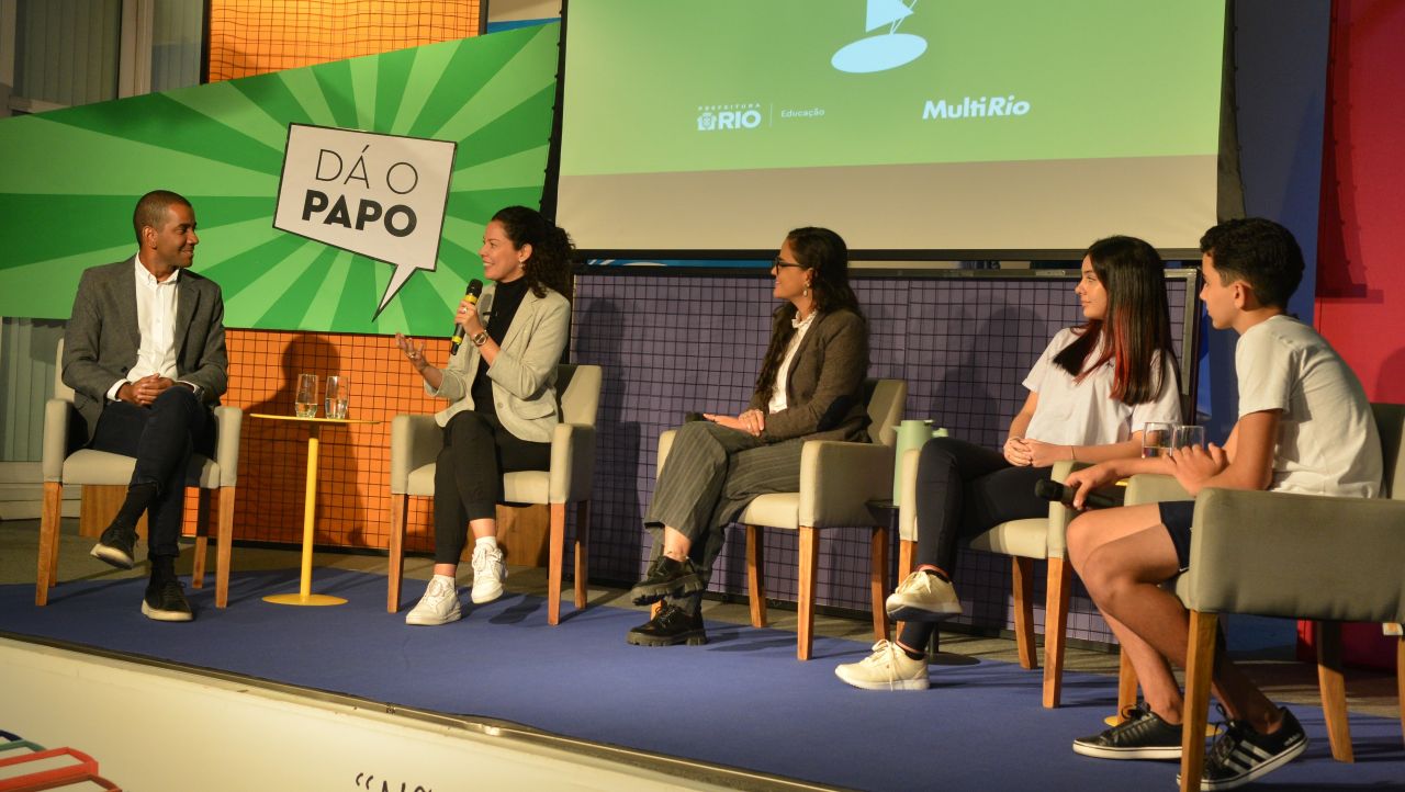 Foto. Cinco pessoas estão sentadas em cadeiras em um palco participando de uma discussão, sendo três adultos e dois estudantes de diferentes gêneros e raças. As cadeiras de cores claras estão dispostas em linha. No fundo, painéis coloridos, um deles com o logotipo do jogo Dá o papo, e um banner para projeção.