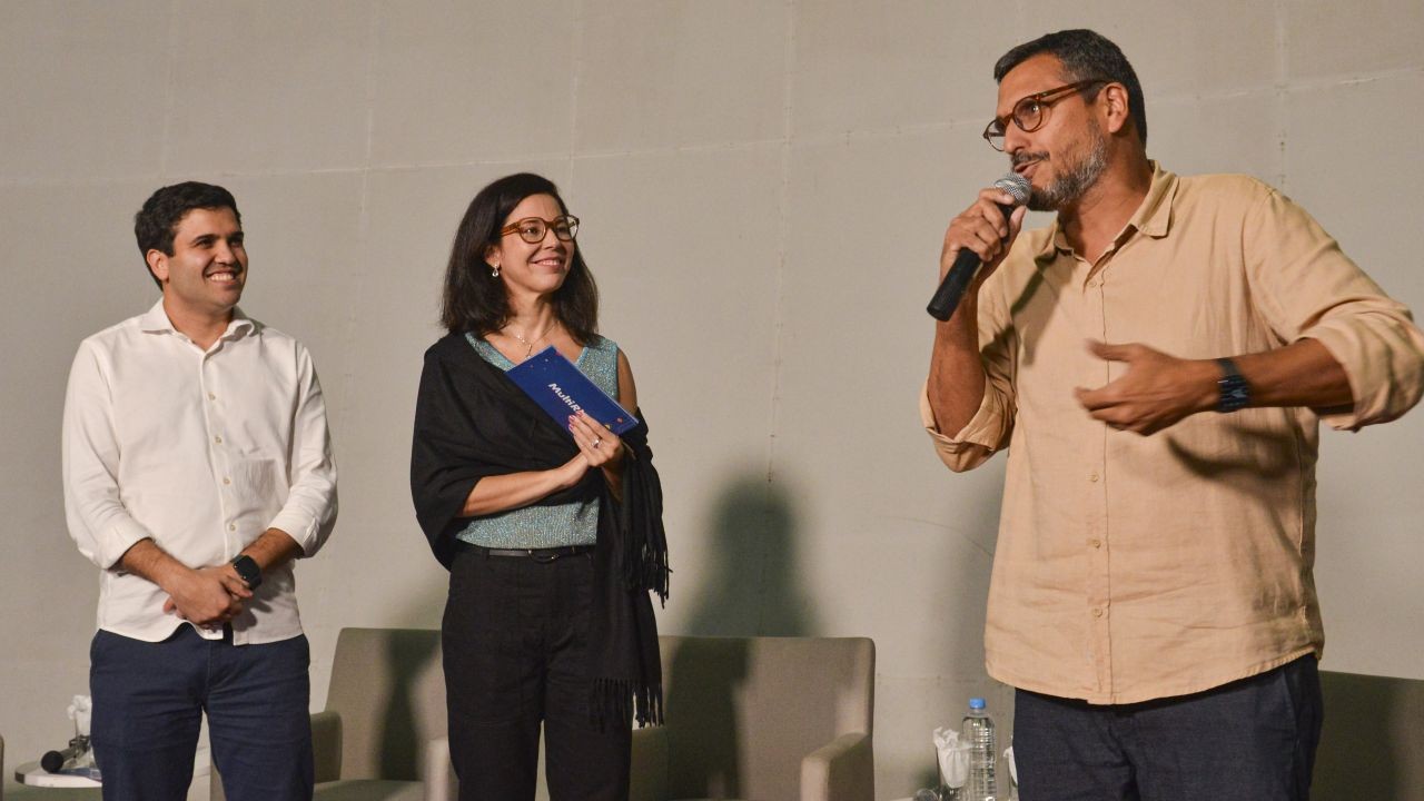 Foto. Um homem com microfone na mão está falando; um homem e uma mulher ao lado dele, também com microfones em mãos, o olham e sorriem.